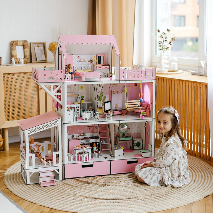 A child sitting in front of a two-story wooden dollhouse that is predominantly white with pink accents. The dollhouse has a balcony and a terrace.