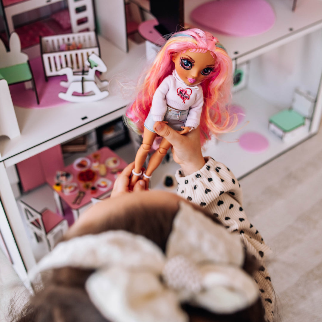 Child holding a LOL doll with pink hair in a playroom with toys and furniture.