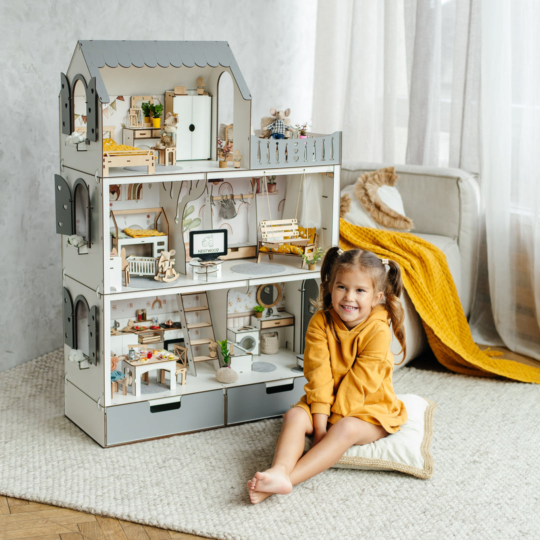 Child sitting on a rug next to a large dollhouse in a room with light gray walls and wooden flooring.