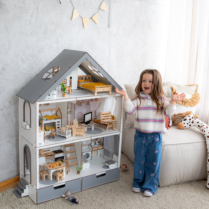 Child standing next to a large dollhouse in a room with a sofa and decorative items.