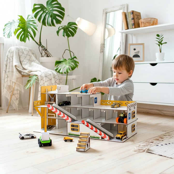 A young child playing with a large toy multi-level parking structure and small construction vehicles in a bright, modern nursery or playroom with light wood floors, white furniture, and large green plants.