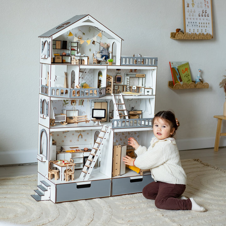 A young child kneels beside a large wooden dollhouse with miniature rooms, furniture, stairs, and ladders in a bright, styled room.