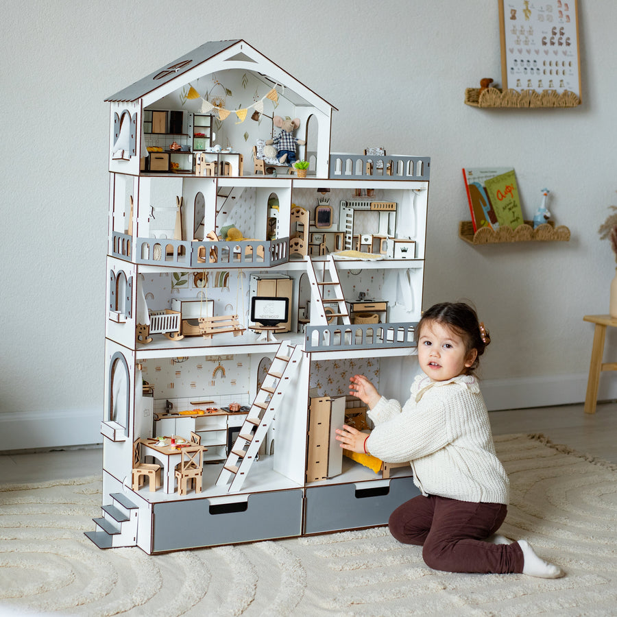 A young child kneels beside a large wooden dollhouse with miniature rooms, furniture, stairs, and ladders in a bright, styled room.