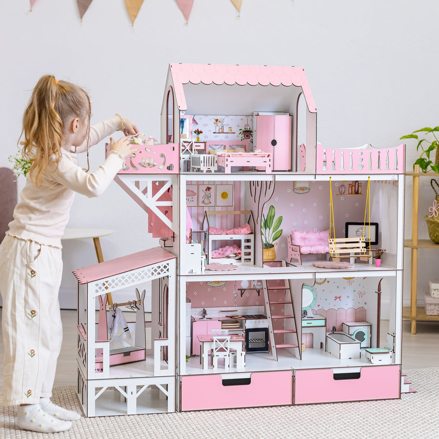 A young girl stands beside a large pink and white dollhouse with miniature furniture and rooms, in a bright, styled bedroom setting.