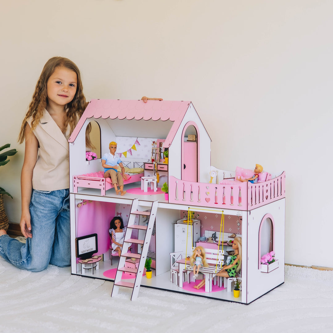 Young girl standing next to a pink dollhouse with furniture and dolls inside.