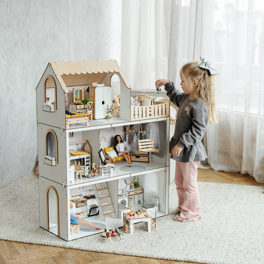 A young girl with blonde hair in a bow stands next to a large, multi-level wooden dollhouse with miniature furniture and accessories, set in a bright room with a textured rug and wood parquet floor.