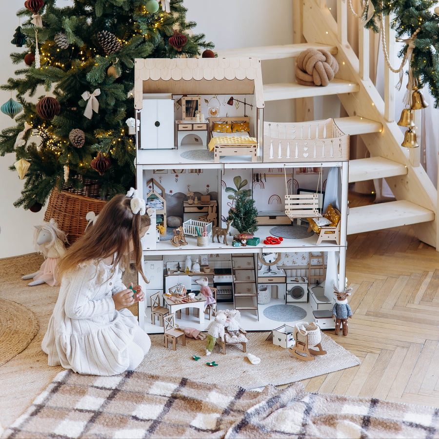 A child in a white dress sits on a plaid rug beside a large, fully furnished dollhouse next to a decorated Christmas tree and a wooden staircase with a green garland.