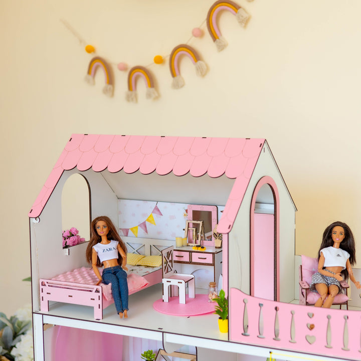 A pink and white dollhouse with two dolls inside, set against a beige wall with a wooden rainbow garland and wall decor.