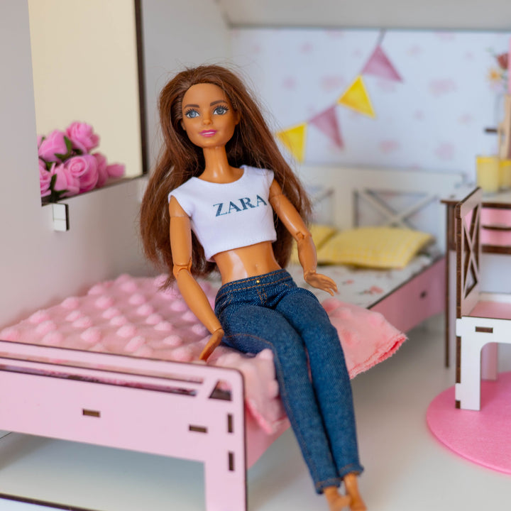 A doll with long brown hair wearing a white crop top with 'ZARA' printed on it and blue jeans, sitting on a pink bed in a miniature doll's bedroom set.
