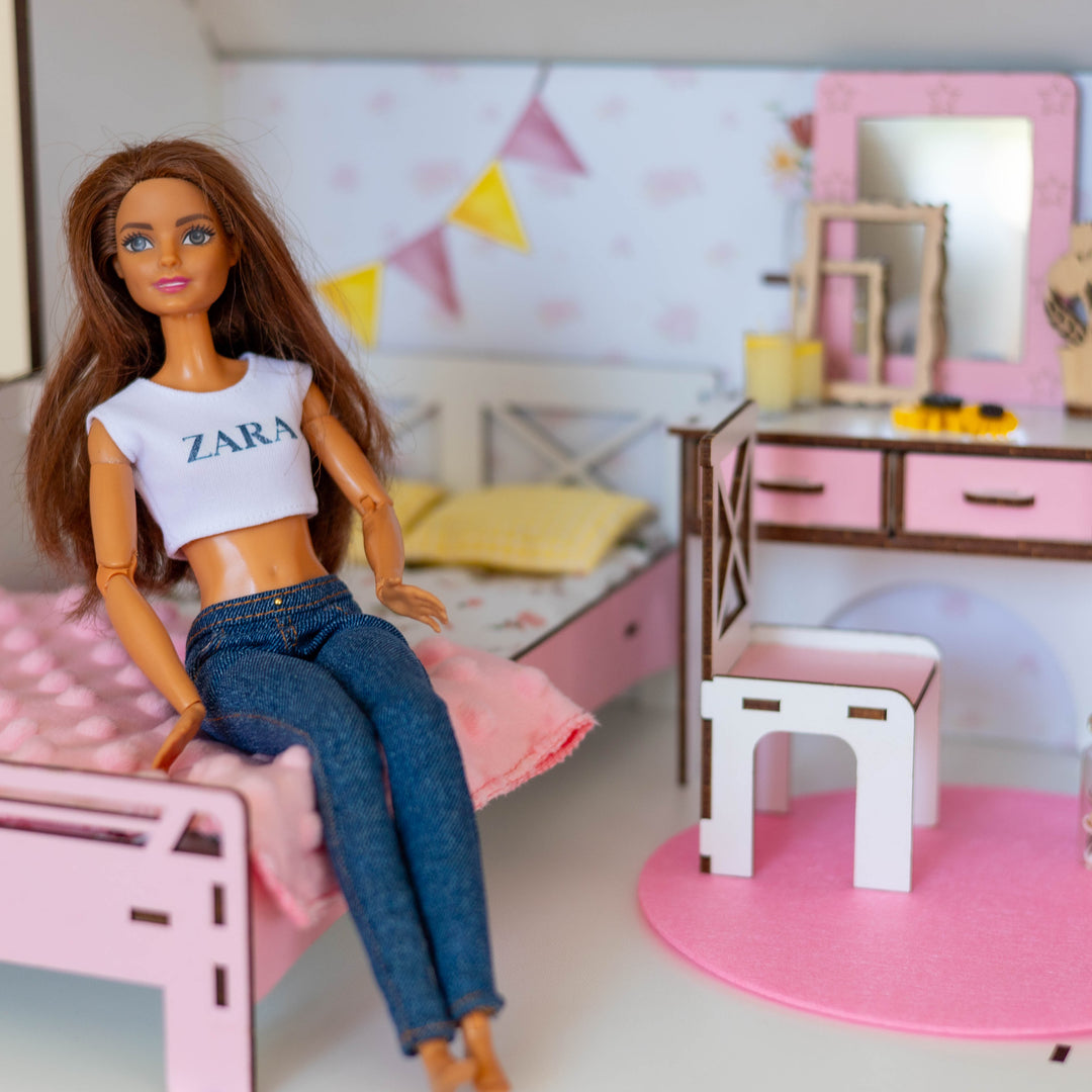 A doll with long brown hair wearing a white crop top with 'ZARA' printed on it and blue jeans, sitting on a pink bed in a miniature doll's bedroom set.