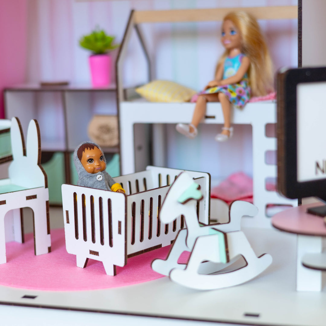 A miniature dollhouse interior scene with a white crib, rocking horse, bunny chair, desk, TV screen reading NESTWOOD, and a doll sitting on a loft bed.
