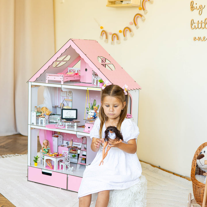 A young girl in a white dress sits on a fluffy white stool in front of a large pink dollhouse, holding a doll in a bright, decorated room with a wooden floor and wall decals.