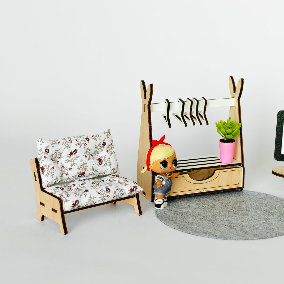A dollhouse miniature furniture set featuring a floral armchair, a wooden clothing rack with hanging clothes, a small potted plant, and a tiny doll standing beside it, all set on a gray circular rug against a white background.
