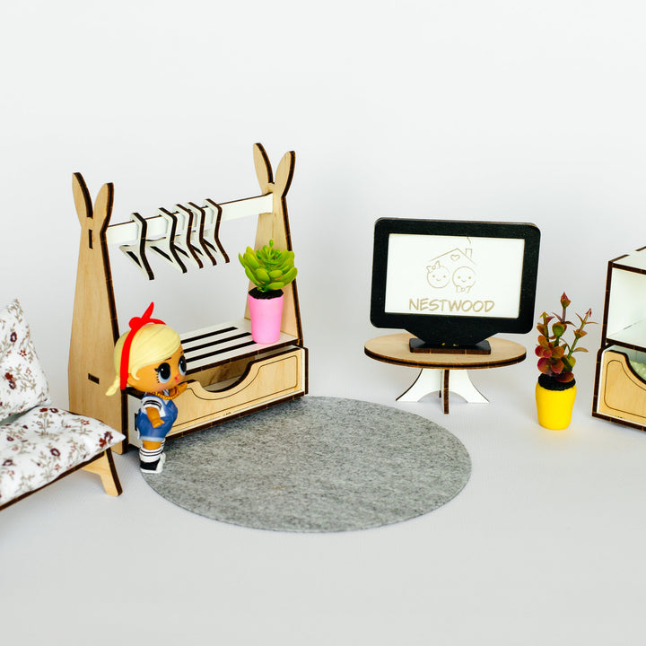 A miniature dollhouse furniture set featuring a wooden A-frame clothing rack with a small plant, a round gray felt rug, a miniature TV stand with a screen displaying 'NESTWOOD', and a small doll, all arranged on a light gray background.
