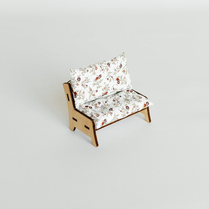 A miniature wooden chair with white cushions featuring a delicate multicolor floral pattern, set against a seamless light gray background.