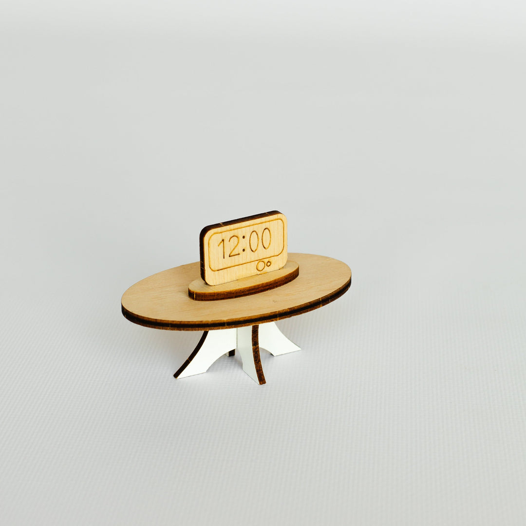 A miniature wooden tabletop clock figurine featuring a round light brown wood base with three small legs and a small rectangular wooden sign on top displaying '12:00'.