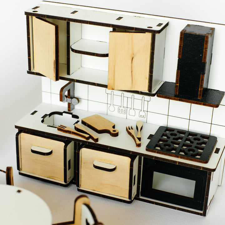 A miniature laser-cut wooden toy kitchen set with light natural wood panels and black accents, including cabinets, drawers, a stove, and utensils, shown on a white background.