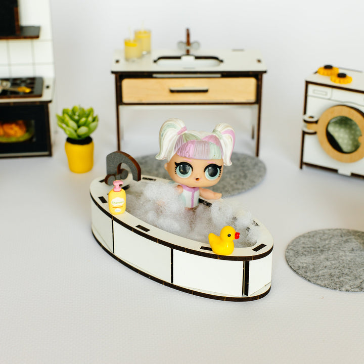A miniature doll with pink hair sits in a small white bathtub filled with faux bubbles, surrounded by tiny bath accessories. The scene is set in a tiny toy room with a vanity, washing machine, and small plant, all photographed on a clean white surface.