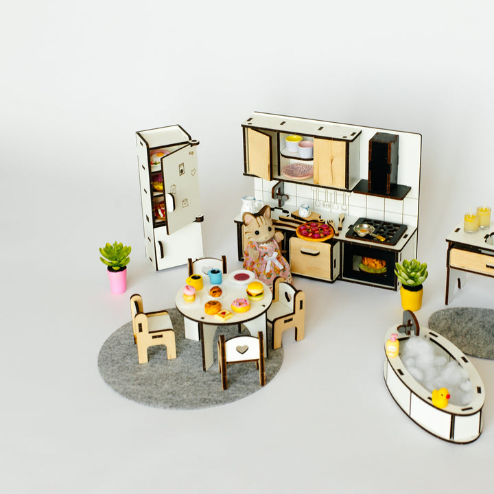 A miniature dollhouse-style kitchen and dining set with a small cat figurine sitting at a round table. The set includes a refrigerator, kitchen cabinets, stove, dining chairs, a round dining table with food, potted plants, and a bathtub, all arranged on a plain light background.
