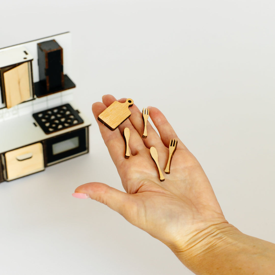 A hand holding several miniature gold-colored wooden kitchen utensils, with a small toy kitchen set visible in the blurred background on a clean white surface.