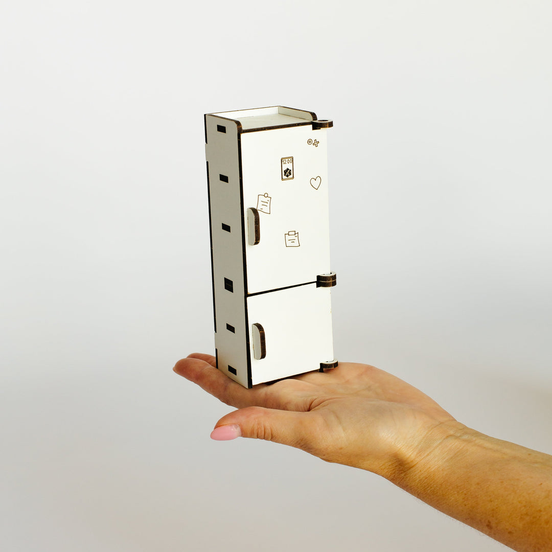 A miniature white laser-cut wooden toy refrigerator held in a person's hand against a plain light gray background.