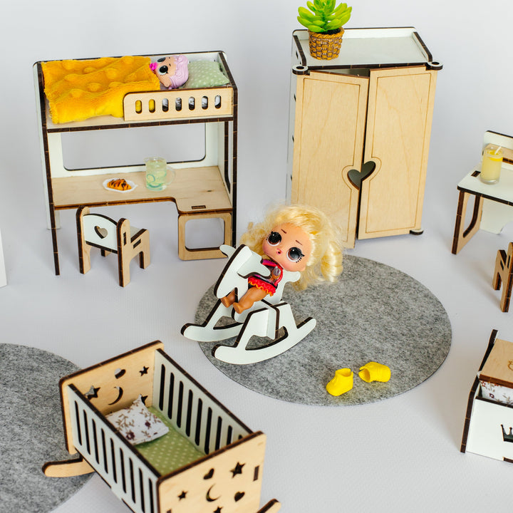A miniature dollhouse nursery set featuring a wooden crib, cabinet, desk, rocking chair with a doll, and other small furniture pieces arranged on gray felt rugs against a white background.