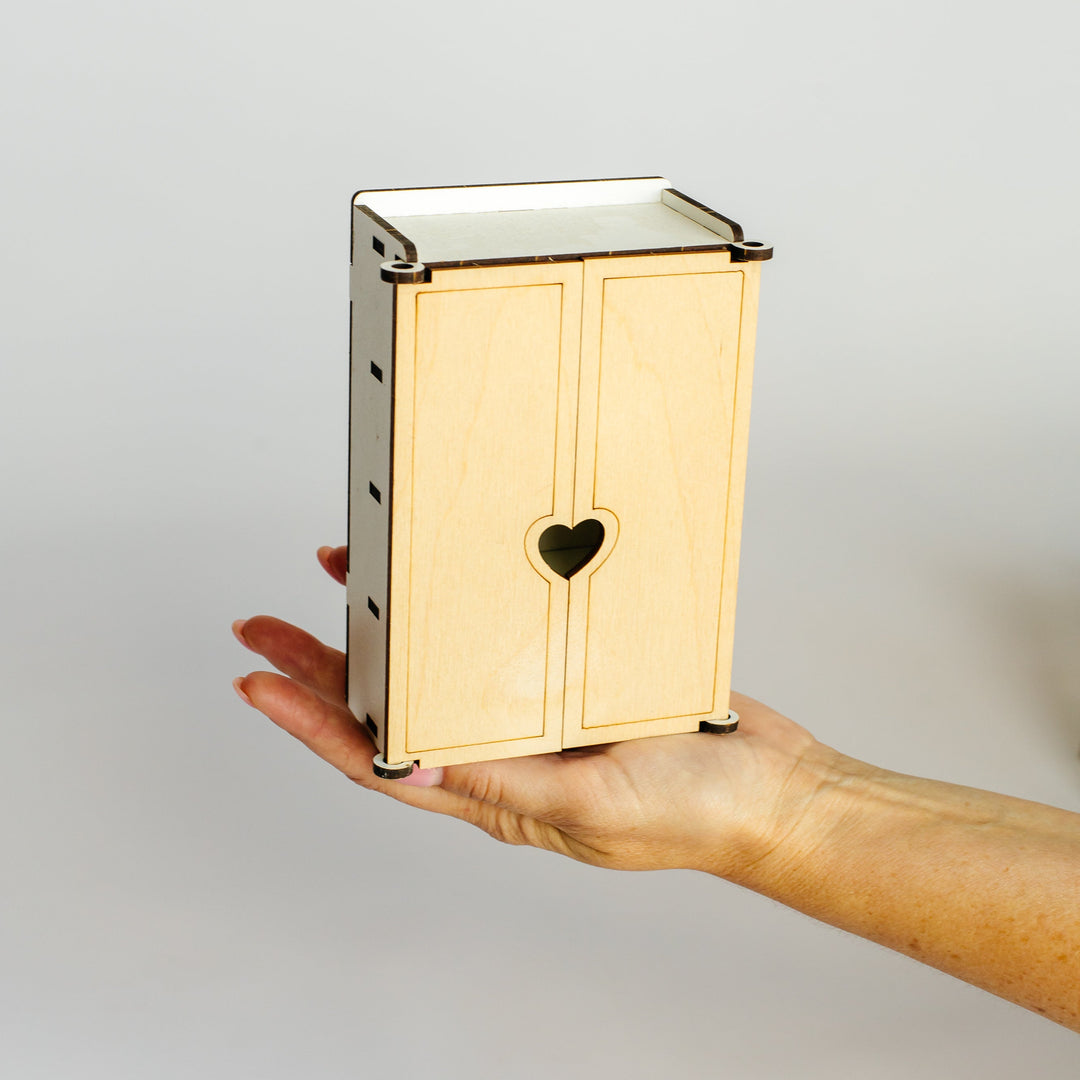 A small wooden cabinet with two doors and a heart-shaped cutout in the center, held in a person's hand against a plain light gray background.