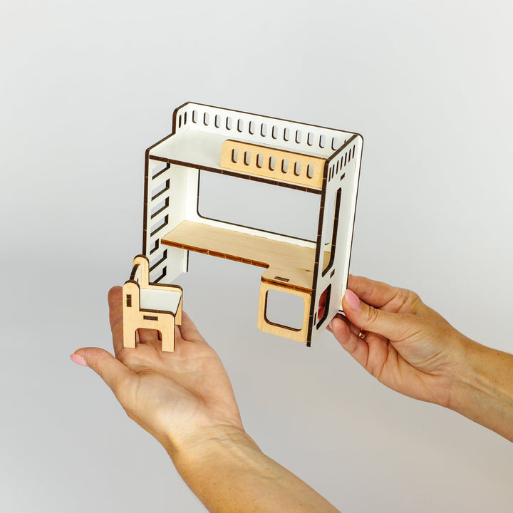 A person's hands holding a miniature laser-cut wooden desk and chair model against a plain light gray background.
