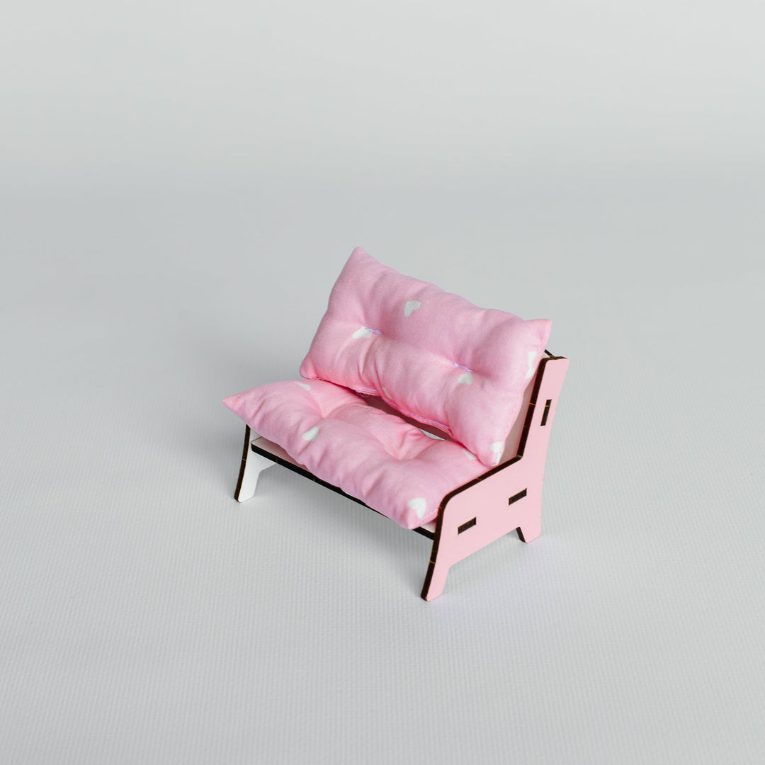 A miniature pink futon-style daybed with tufted cushions and a light wood frame, photographed on a seamless light gray studio background.