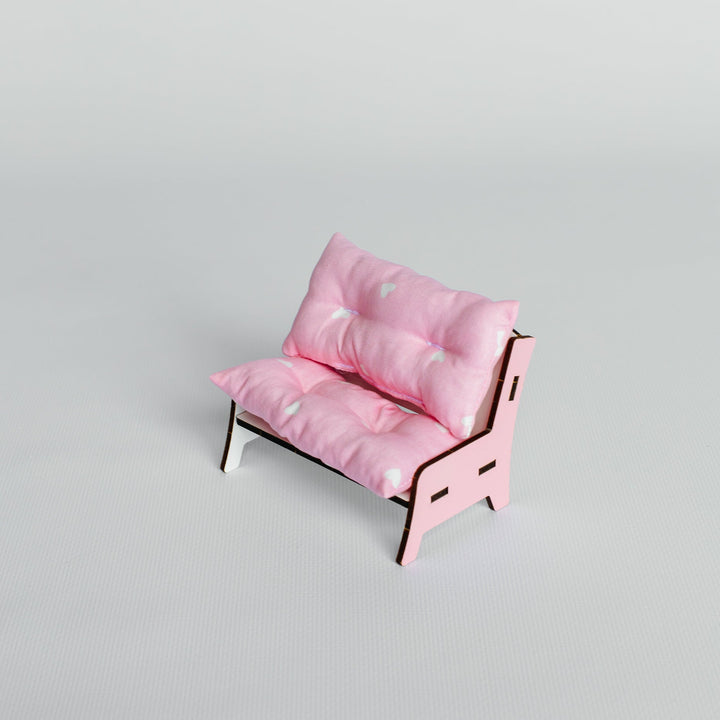 A miniature pink futon-style daybed with tufted cushions and a light wood frame, photographed on a seamless light gray studio background.