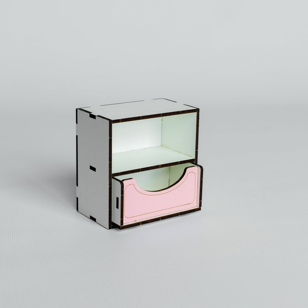 A minimalist two-tier storage organizer made of laser-cut light wood, featuring a white top tier and a pink bottom drawer with a dark metal frame, set against a solid light gray background.