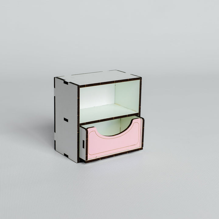 A minimalist two-tier storage organizer made of laser-cut light wood, featuring a white top tier and a pink bottom drawer with a dark metal frame, set against a solid light gray background.