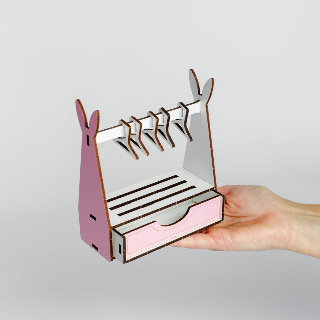 A hand holding a small pink and white wooden rack or organizer with curved side supports and horizontal slats, shown against a light gray studio background.