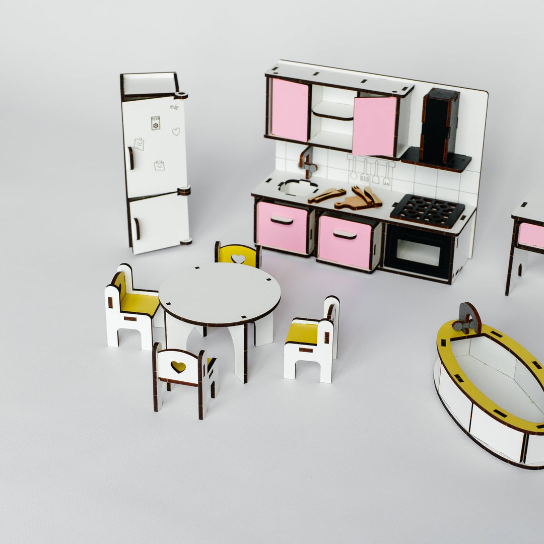 A miniature toy kitchen and dining set displayed on a light gray background. The set includes a small refrigerator, dining table, chairs with yellow seats, a play kitchen with pink cabinets, and a yellow-and-black oval accessory piece.