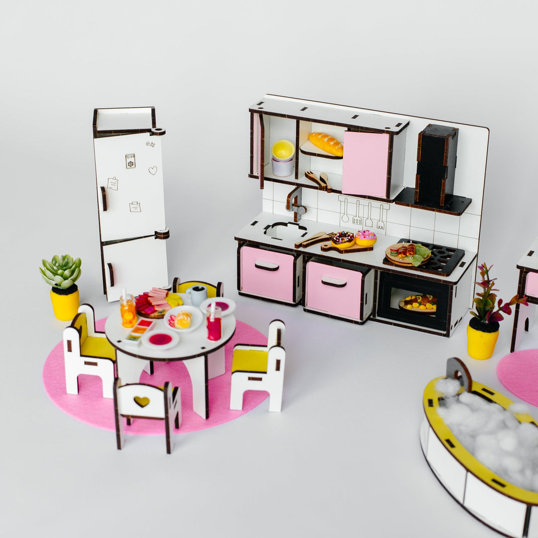 A miniature dollhouse-style kitchen and dining set with pink cabinets, a white refrigerator, a round dining table with chairs, small potted plants, and a bathtub, photographed on a clean light gray background.