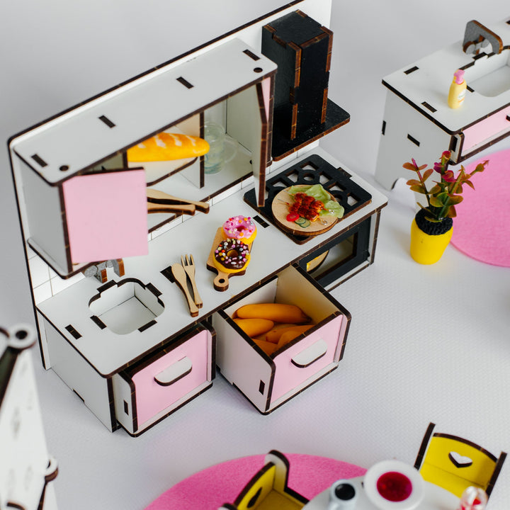A miniature toy kitchen and dining set made from layered wood sheets, featuring a white and pink kitchen with a stove, oven, sink, and drawers, alongside a small round dining table with chairs and pink placemats on a white background.