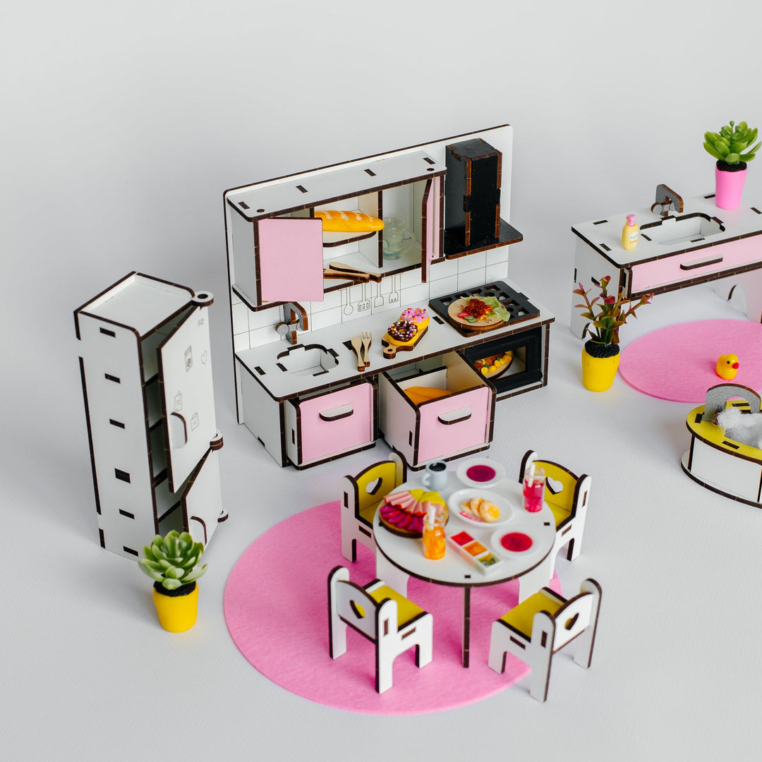 A miniature toy kitchen set with pink and white cabinets, a dining table with chairs, small potted plants, and round pink mats, arranged on a light gray background.