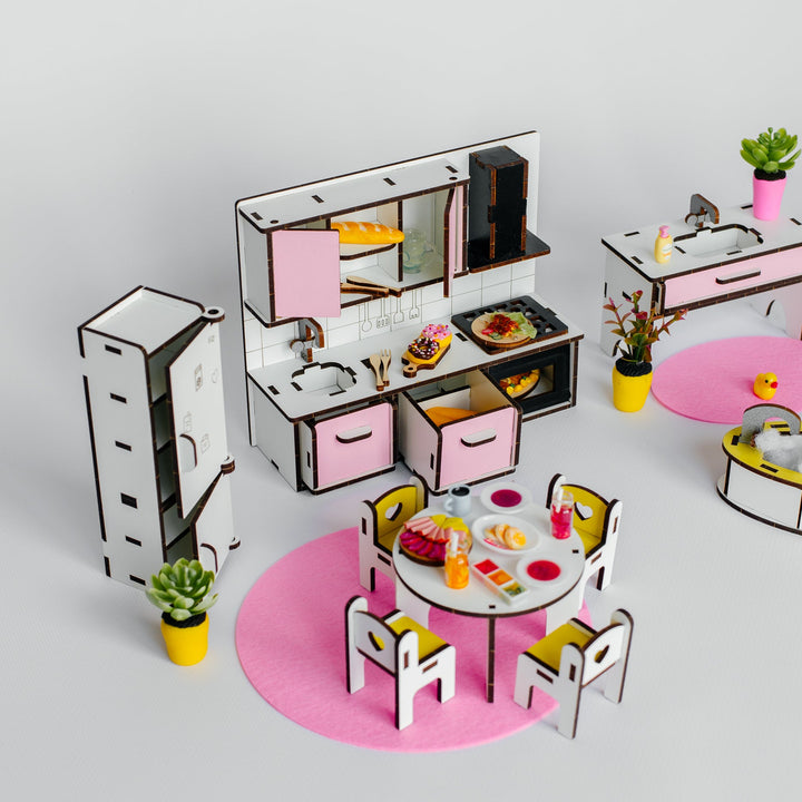 A miniature toy kitchen set with pink and white cabinets, a dining table with chairs, small potted plants, and round pink mats, arranged on a light gray background.