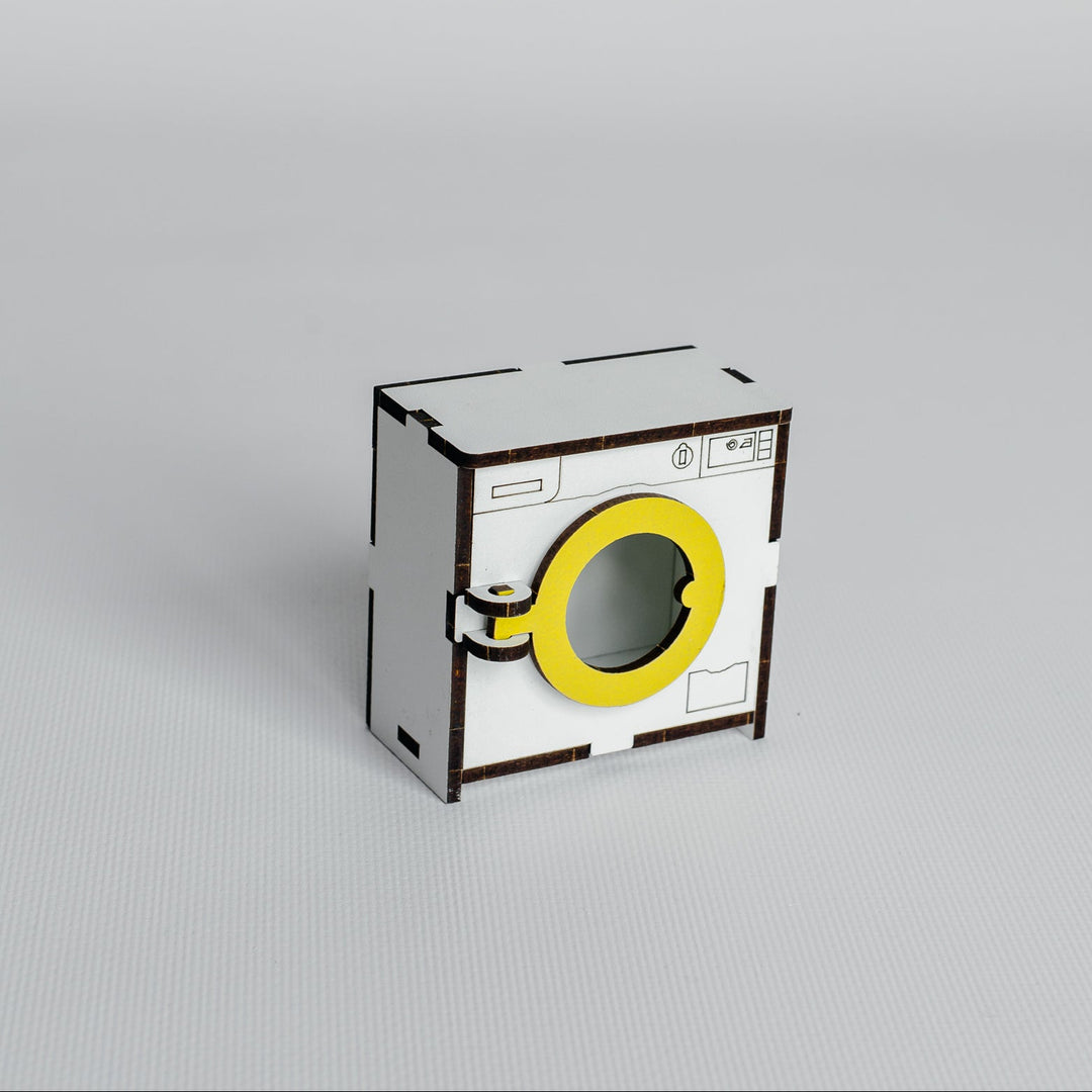 A miniature cardboard model of a washing machine, featuring a white body, a bright yellow circular door, and black printed details, set against a seamless light gray background.