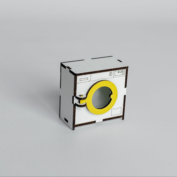 A miniature cardboard model of a washing machine, featuring a white body, a bright yellow circular door, and black printed details, set against a seamless light gray background.