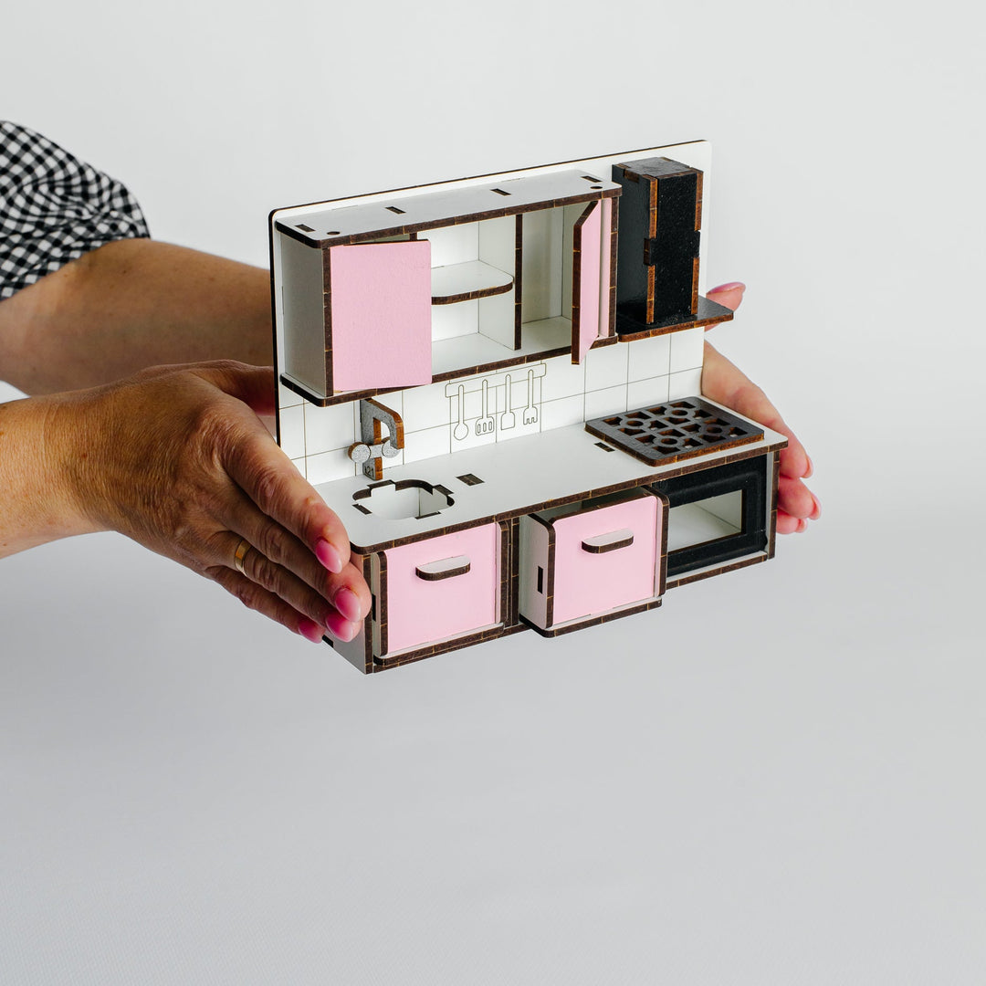 A miniature kitchen playset with pink cabinets, a white countertop, and black stove and oven details, held by two hands against a light gray background.