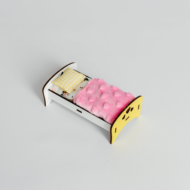 A miniature wooden doll's bed with a white frame, yellow headboard and footboard, a pink tufted blanket, and a yellow-and-white checkered pillow, shown on a light gray background.