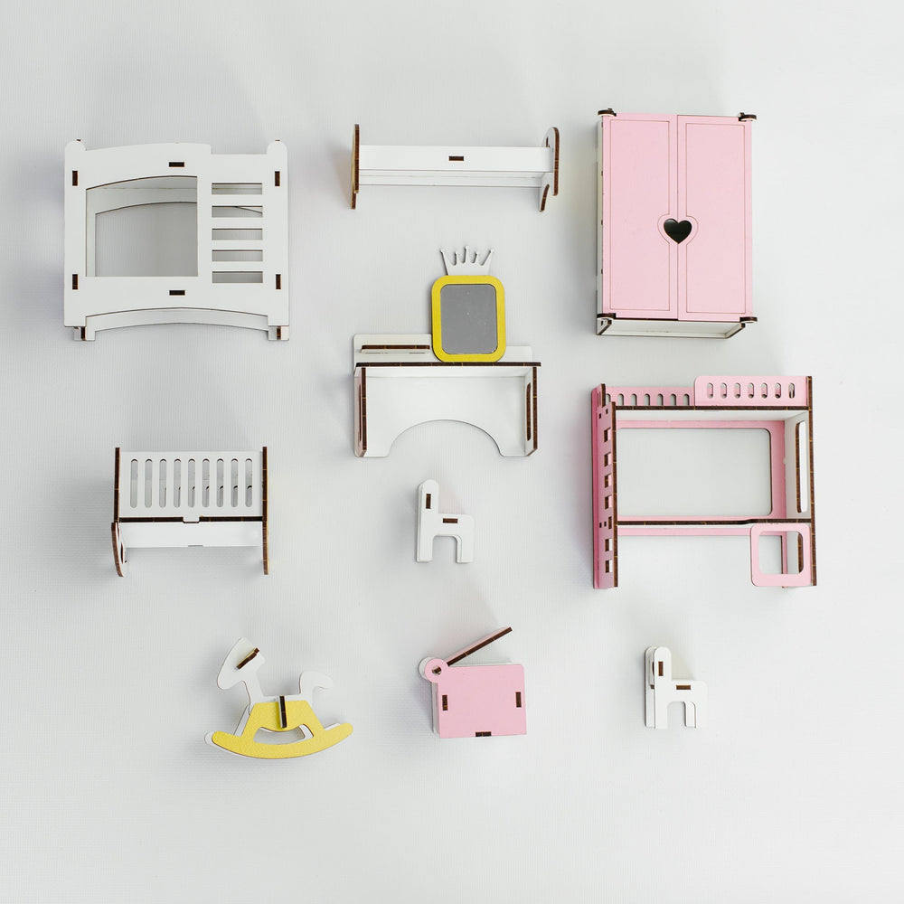 A set of miniature wooden dollhouse furniture pieces and accessories arranged neatly on a light gray background. The collection includes a white crib, a pink wardrobe with a heart cutout, a white dresser with a yellow TV, a pink bed frame, a yellow rocking horse, and other small matching pieces.