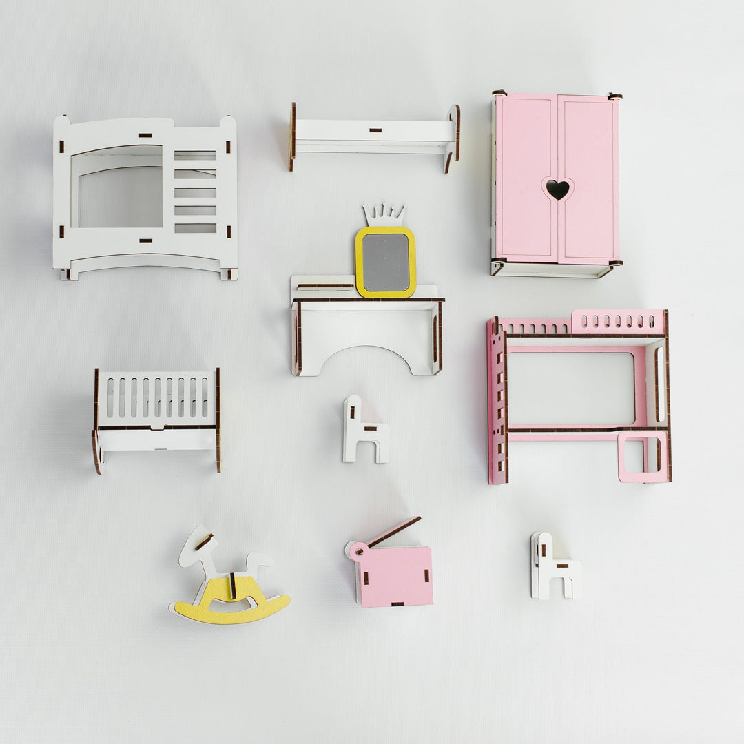 A set of miniature wooden dollhouse furniture pieces and accessories arranged neatly on a light gray background. The collection includes a white crib, a pink wardrobe with a heart cutout, a white dresser with a yellow TV, a pink bed frame, a yellow rocking horse, and other small matching pieces.