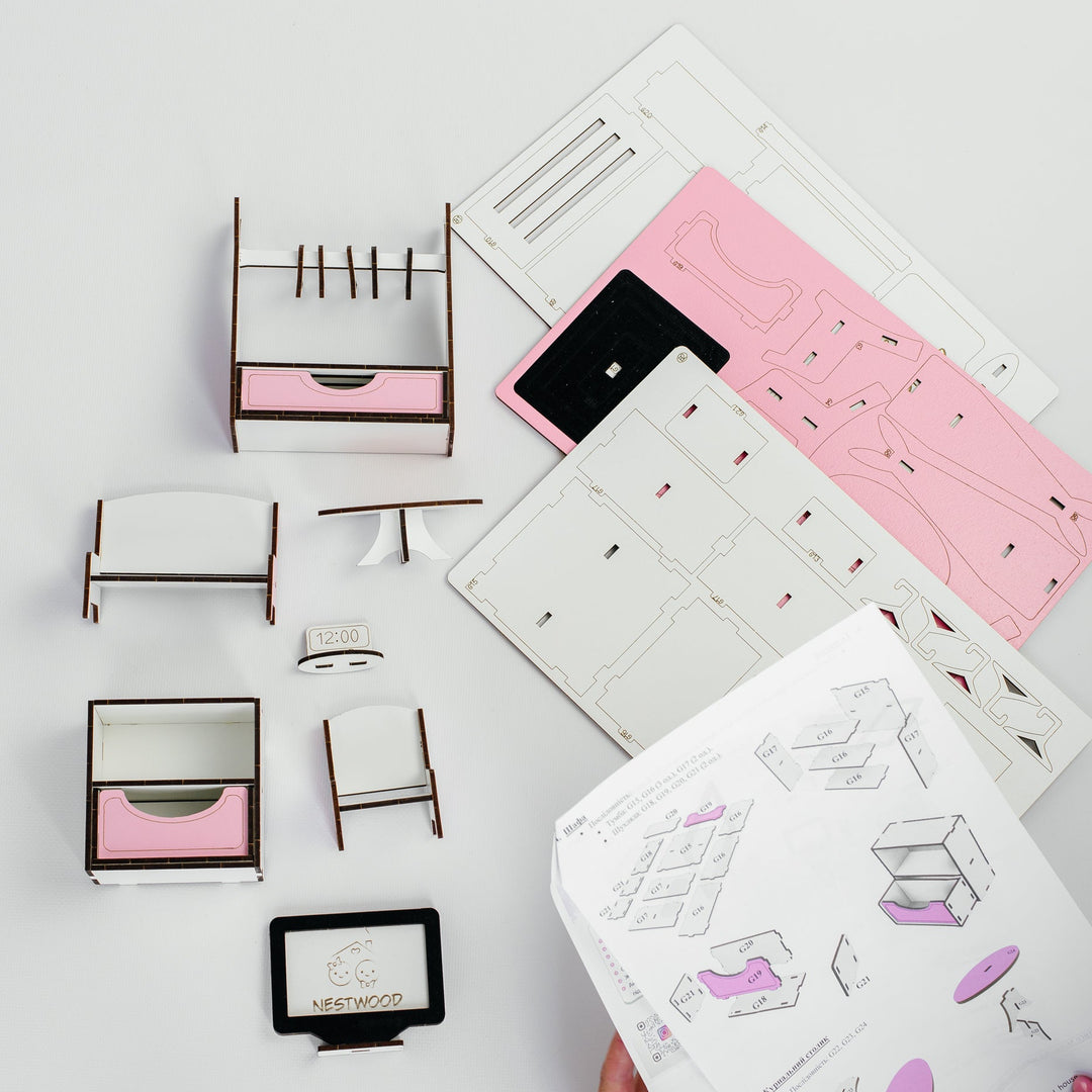 A hand holds a printed assembly manual while several small furniture pieces and flat template sheets are arranged on a light gray surface. The set appears to be a DIY wooden storage cabinet or organizer kit, with pink drawer fronts and white panels.