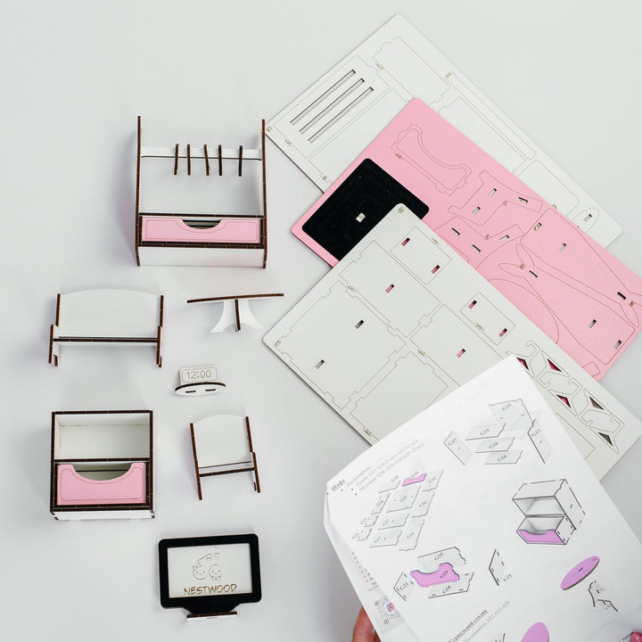 A hand holds a printed assembly manual while several small furniture pieces and flat template sheets are arranged on a light gray surface. The set appears to be a DIY wooden storage cabinet or organizer kit, with pink drawer fronts and white panels.