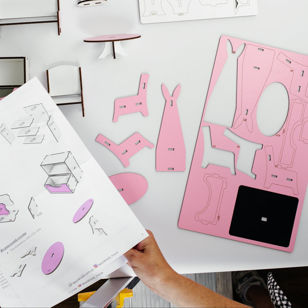 A top-down view of a miniature furniture DIY assembly kit on a white table. The kit includes pink and white wooden pieces for a dresser, shelves, stools, and chairs, along with printed assembly instructions and templates.