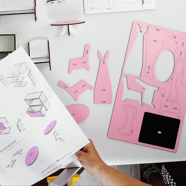 A top-down view of a miniature furniture DIY assembly kit on a white table. The kit includes pink and white wooden pieces for a dresser, shelves, stools, and chairs, along with printed assembly instructions and templates.