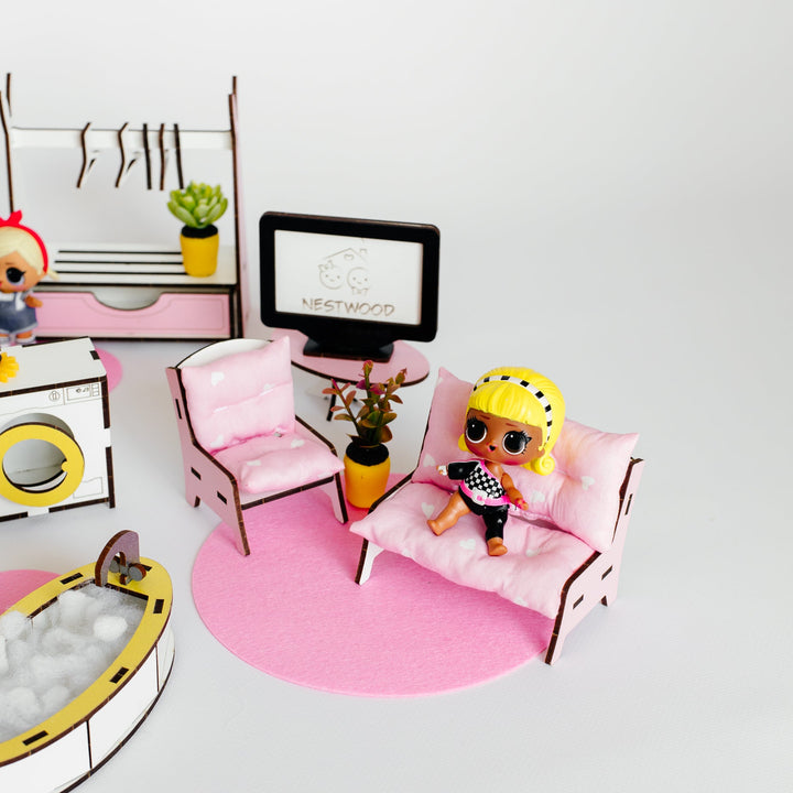 A miniature dollhouse living room scene featuring a pink sofa with a small doll wearing a yellow helmet, a pink chair, a TV, a clothing rack, and a pink circular rug on a white background.