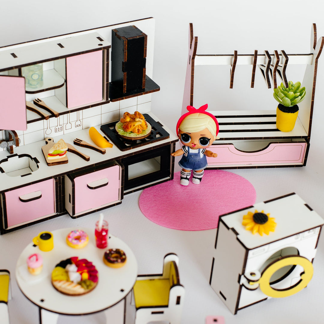 A miniature dollhouse kitchen and room set with pink and white furniture, a small doll standing on a pink rug, a dining table with toy food, a clothing rack, and a washing machine, all arranged on a white surface against a white background.